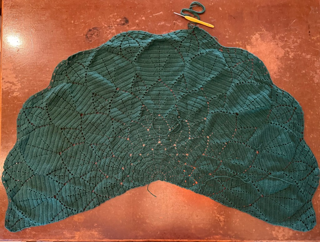 An almost complete shawl crocheted from viridian green cotton-wool blended yarn with the crochet hook at the top of the image. It's laying on a weathered brown wood desktop 